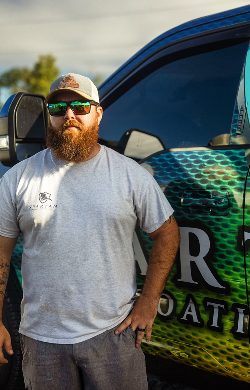 Headshot 1 Man with a baseball cap and shades in a grey shirt standing next to a truck.